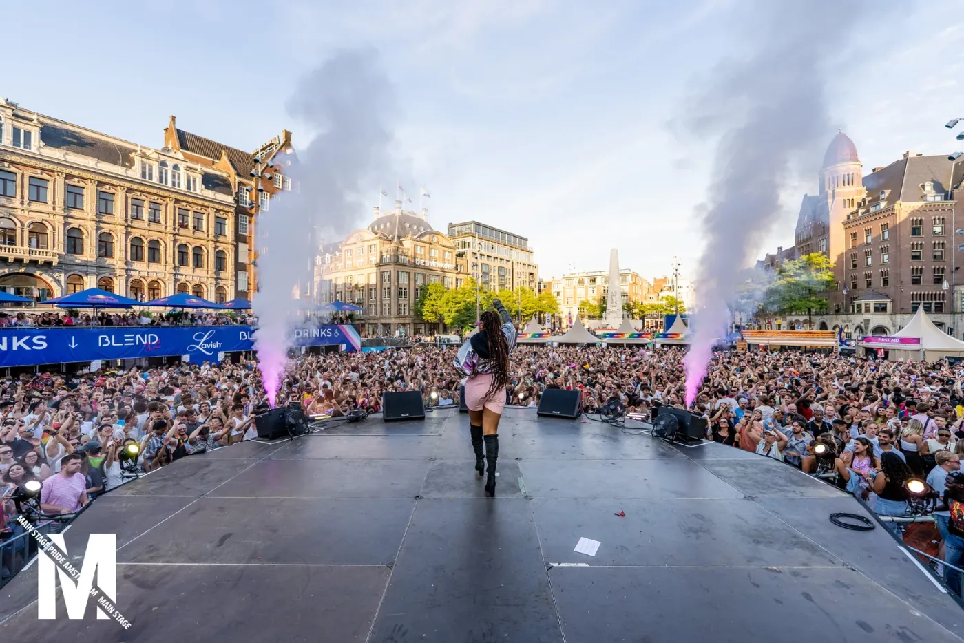 A performer on the MainStage Pride stage between two smoke jets, with a huge crowd on Dam Square