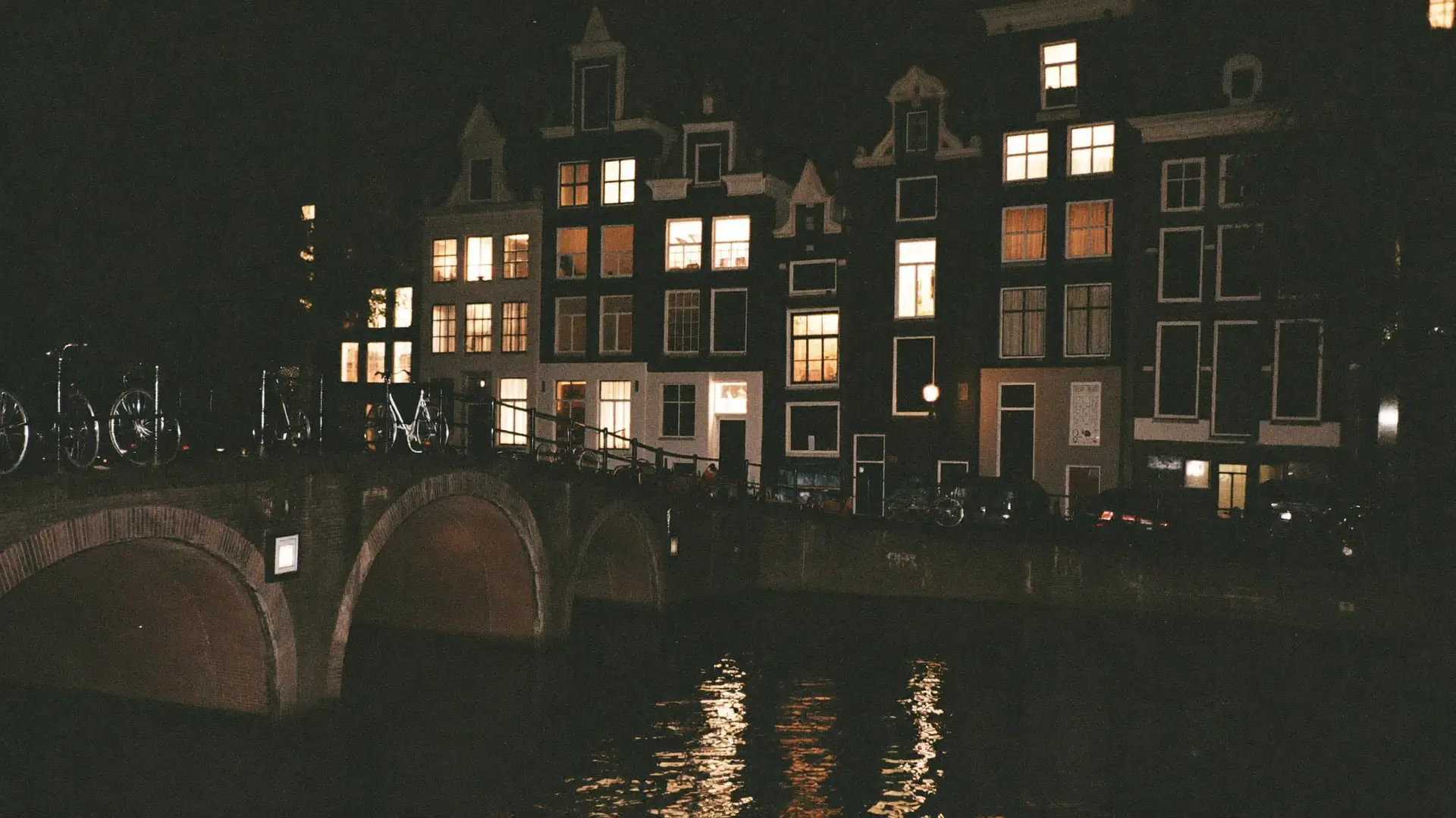 Een Amsterdamse gracht bij nacht na een regenbui, met verlichte grachtenpanden en fietsen op een brug