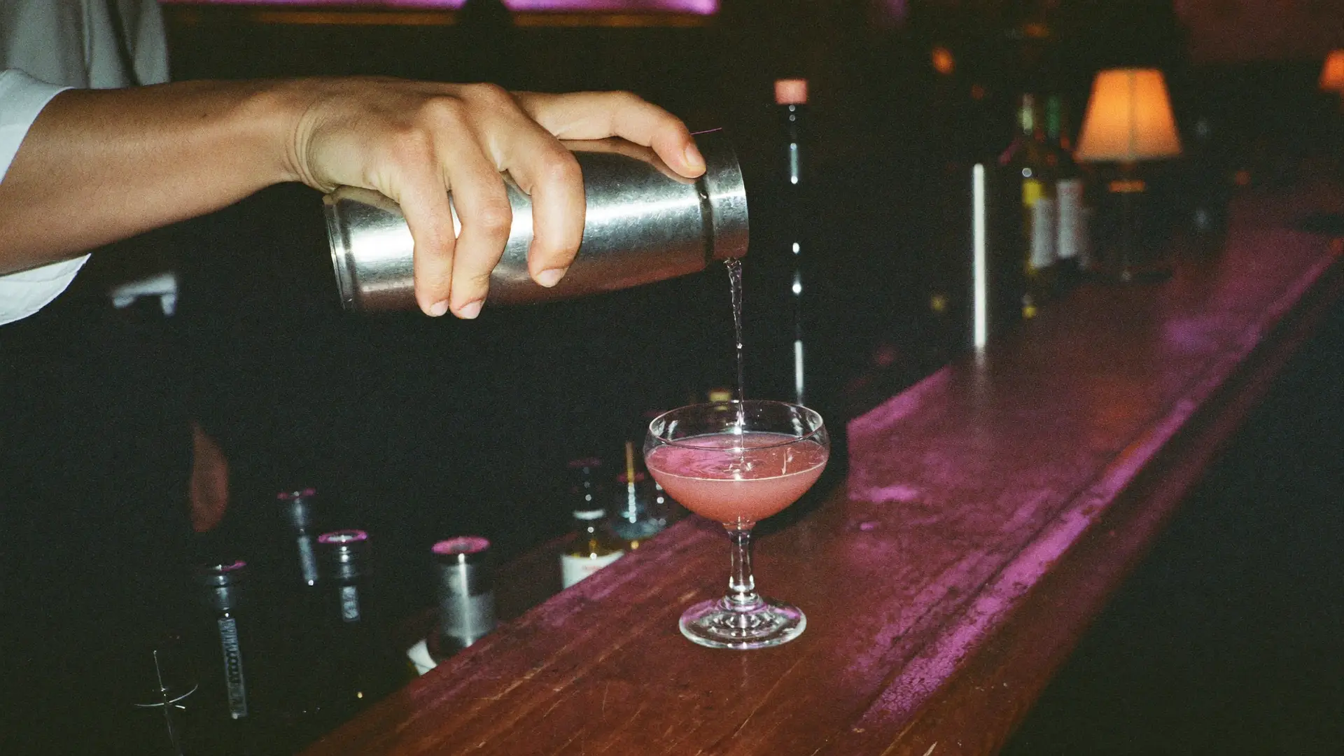 Een roze cocktail wordt uit een shaker in een coupeglas geschonken op een houten bar