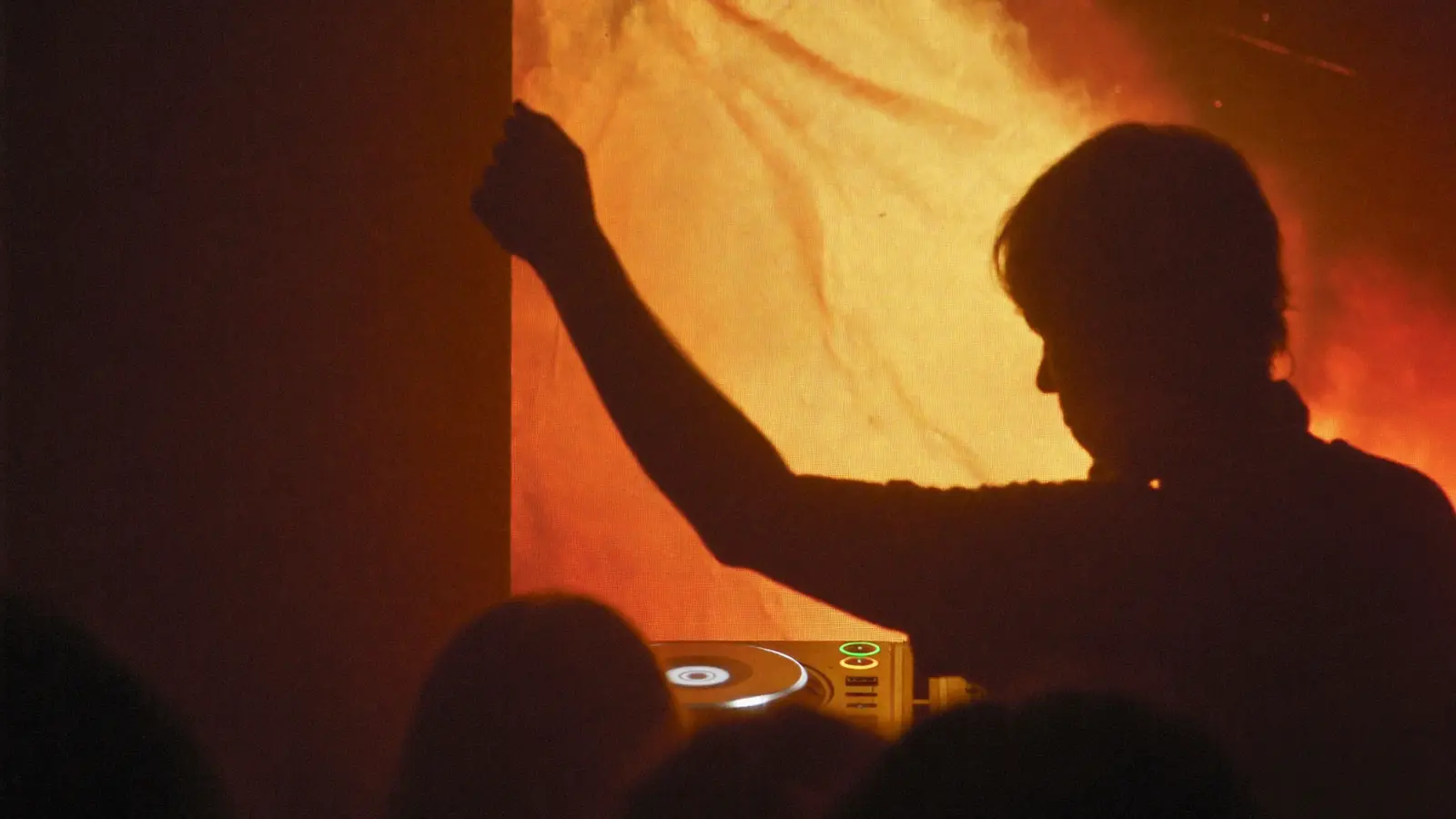 Een DJ in silhouet met een arm omhoog tegen oranje licht