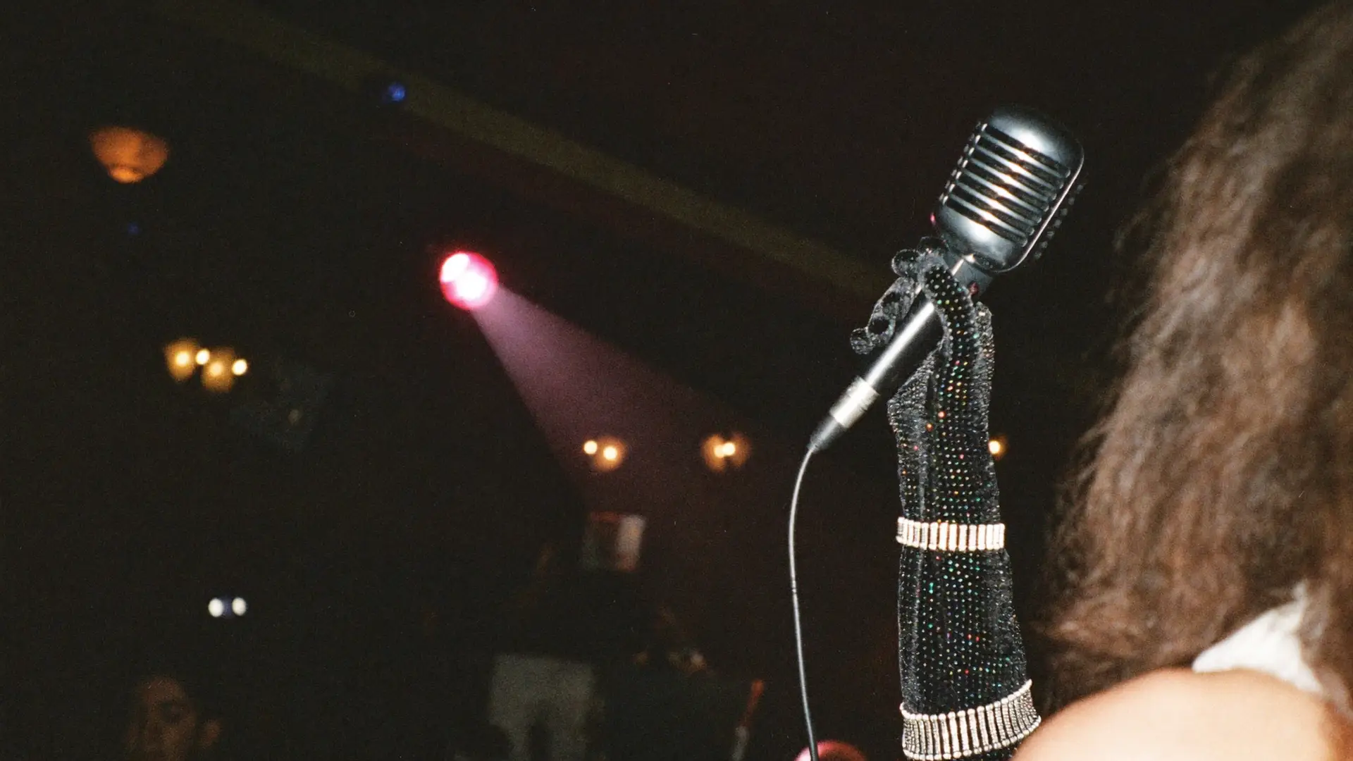 A hand in a glitter glove holding a vintage microphone in a pink spotlight