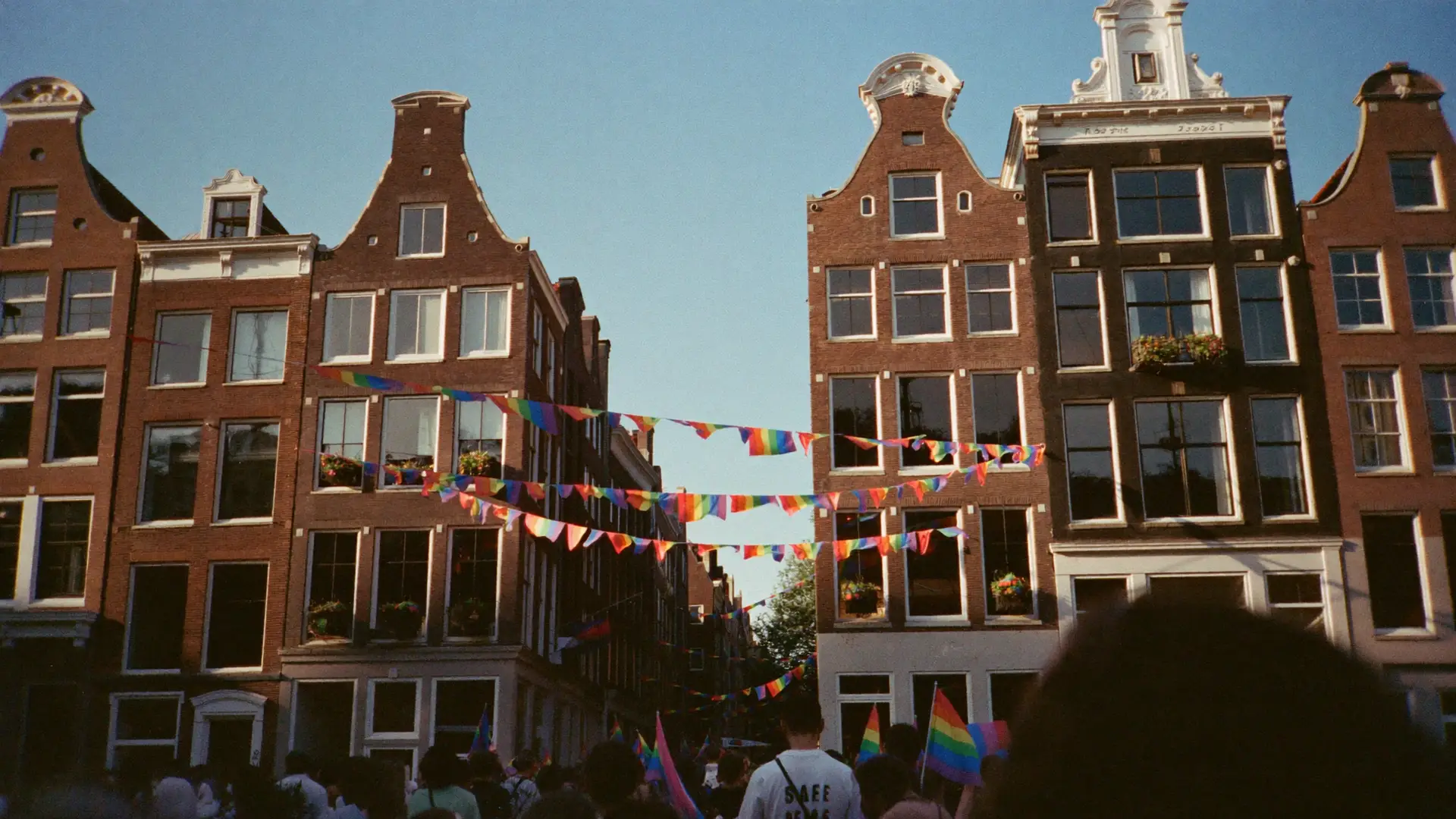 Regenboogvlaggetjes tussen Amsterdamse grachtenpanden boven een feestende menigte