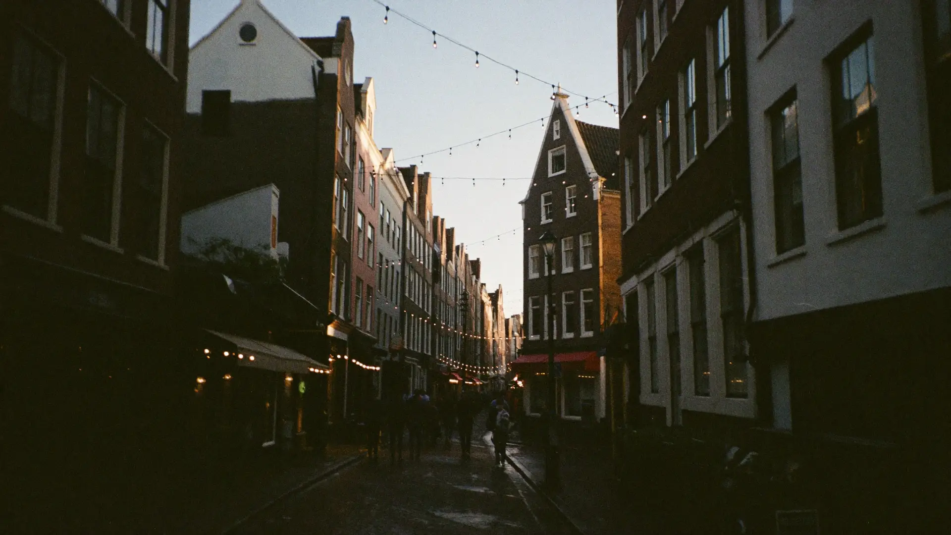 A narrow old Amsterdam street at dusk, with string lights overhead and people heading out